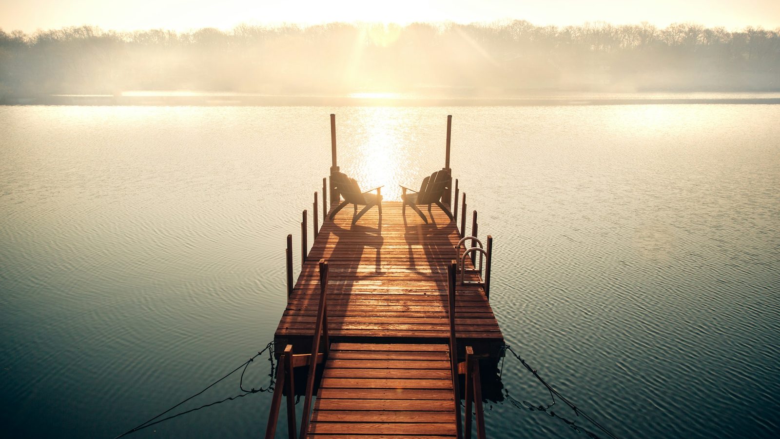 Wooden lake pier with two empty chairs, a swim ladder and calm water at sunrise